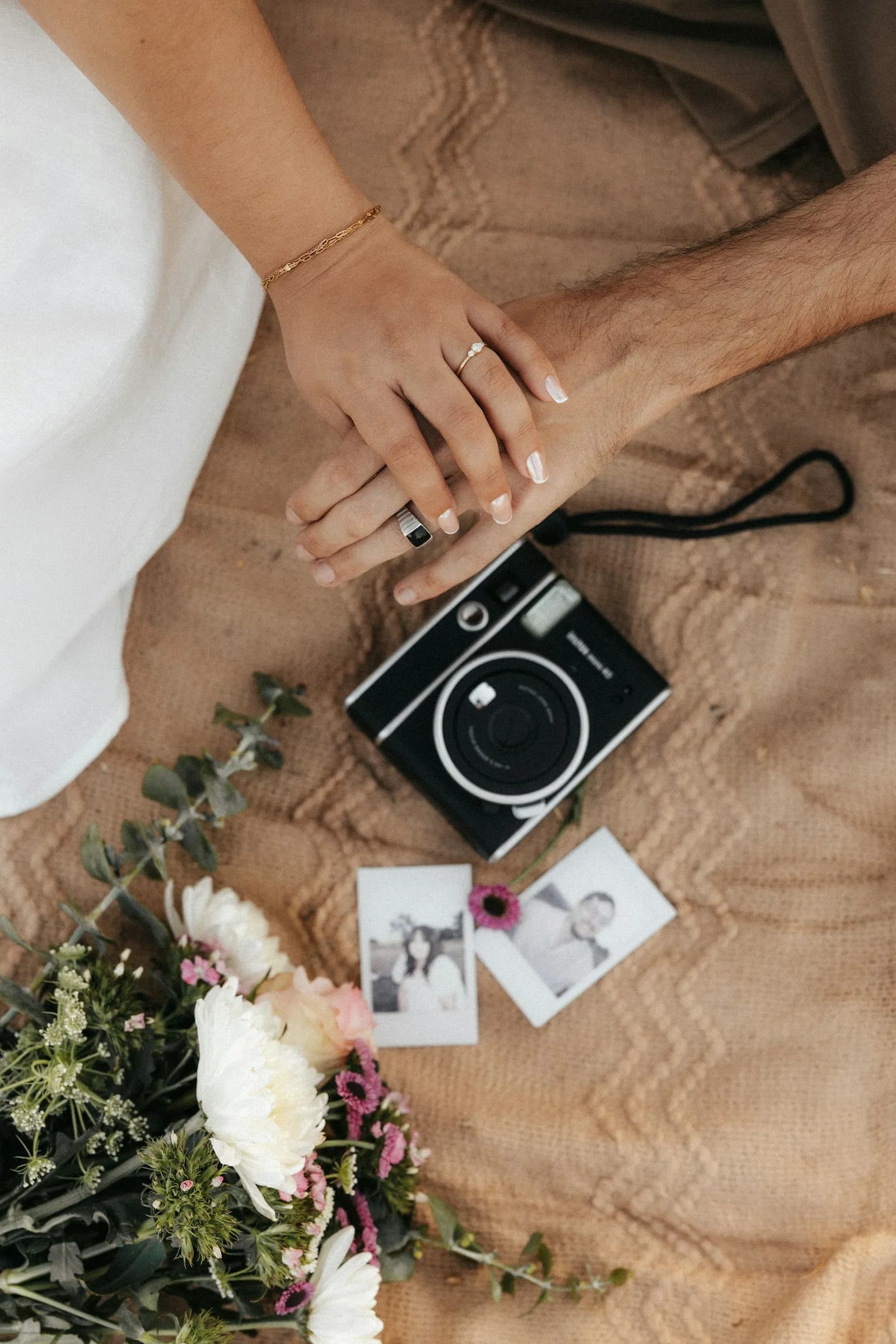 A couple's hands resting beside an instant camera, flowers and two printed photos